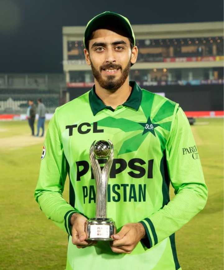 Arafat Minhas in a green Pakistan jersey holding a silver Player‑of‑the‑Match trophy on a stadium field at night