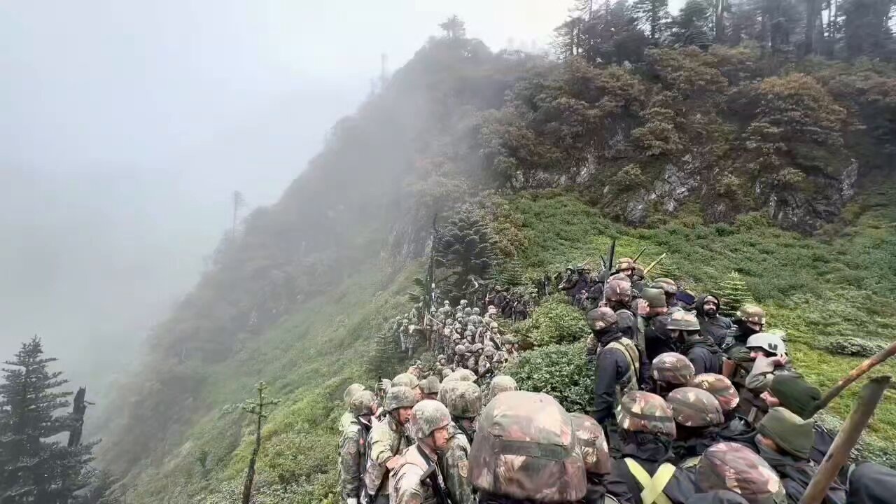 Large group of soldiers in camouflage uniforms and helmets climbing a steep, foggy mountainside with rocky terrain and mist