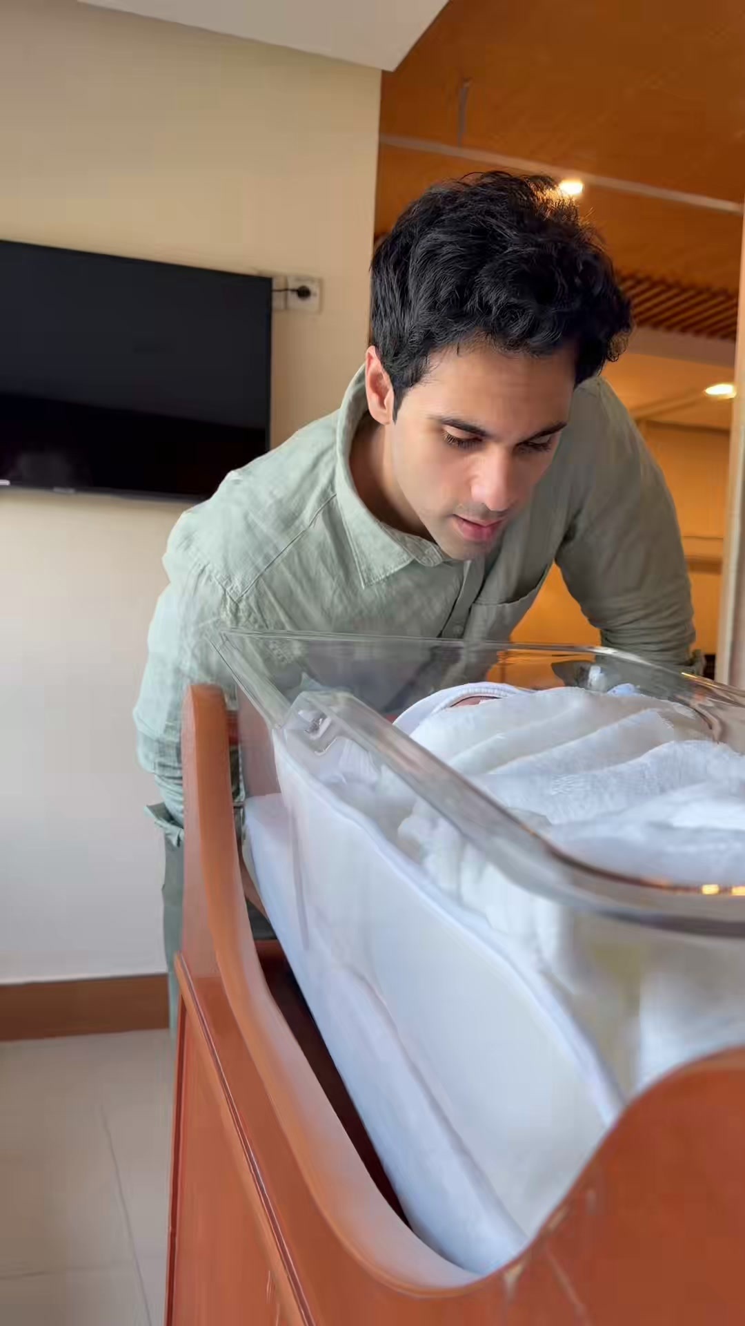 Man in light green shirt leaning over a clear acrylic baby bassinet with a wrapped newborn, TV visible in background