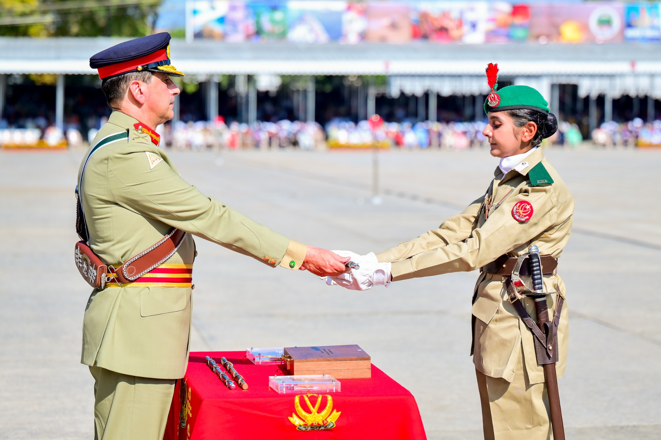 Two military personnel in dress uniforms during a formal ceremony at the Pakistan Military Academy; a man in a tan uniform with peaked cap hands a sword to a woman in a tan uniform wearing a green beret with a red plume, standing beside a red‑cloth‑covered table with ceremonial items under a bright sky