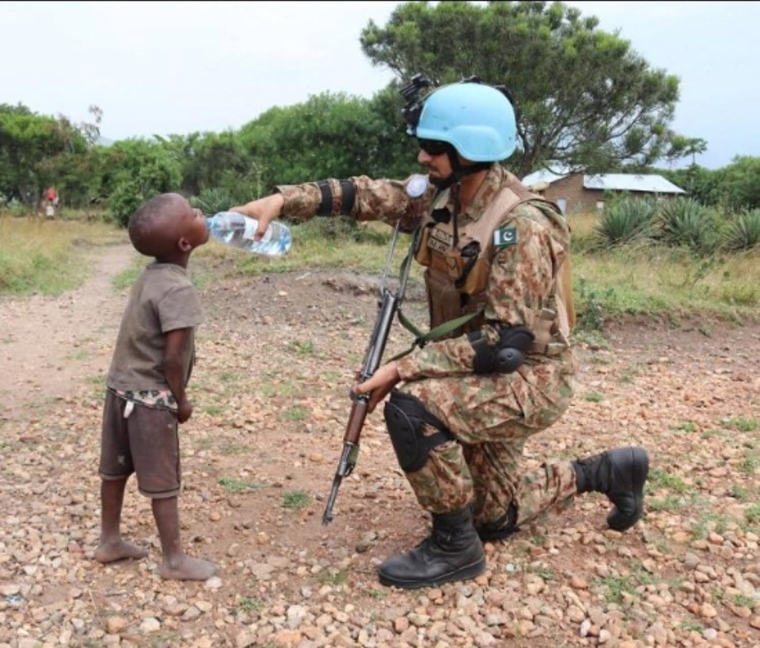 Pakistani soldier in camouflage kneeling and giving water to a barefoot child in a rocky area