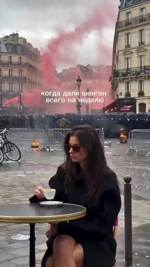 Woman with sunglasses sipping coffee at a Paris café while red smoke and police barriers fill the street behind her