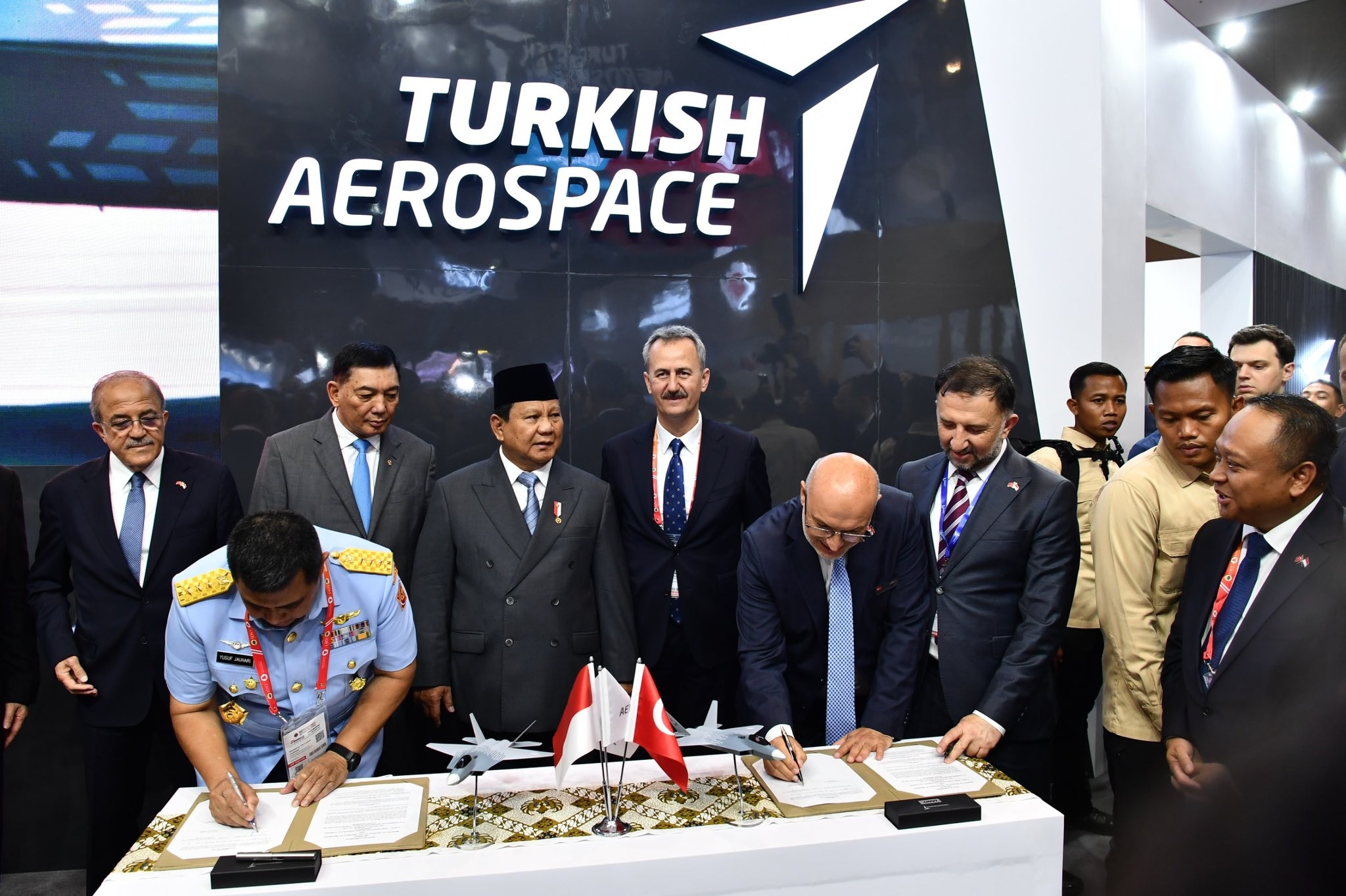 Men in formal attire at a signing ceremony with a backdrop reading 'TURKISH AEROSPACE'; two small flags of Indonesia and Turkey on the table