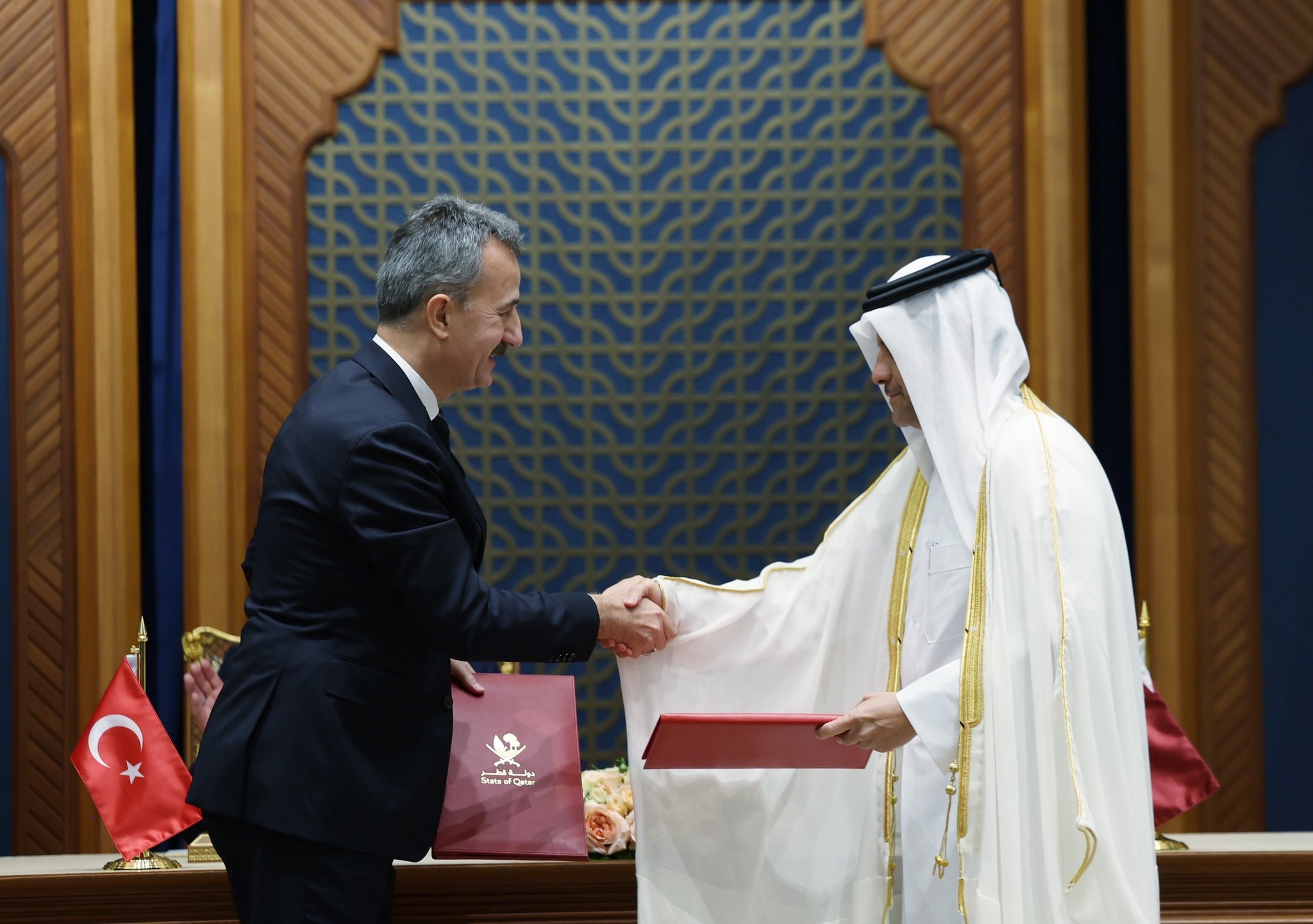 Two men shaking hands, one in a dark business suit and the other in traditional Qatari attire, with Turkish and Qatari flags visible in the background