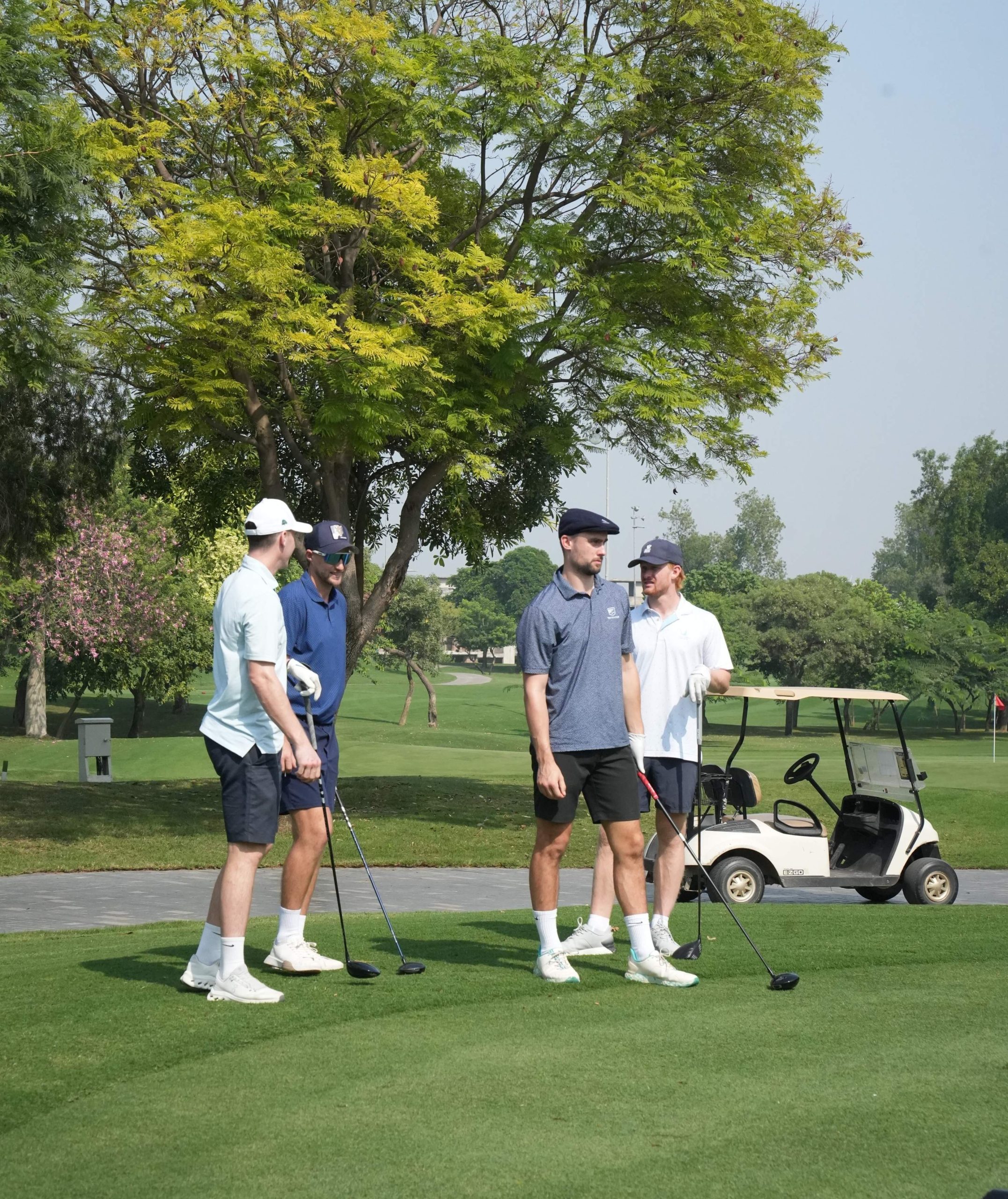 Four men in golf attire standing on a green course beside a white EZGO golf cart, with trees and clear sky in the background