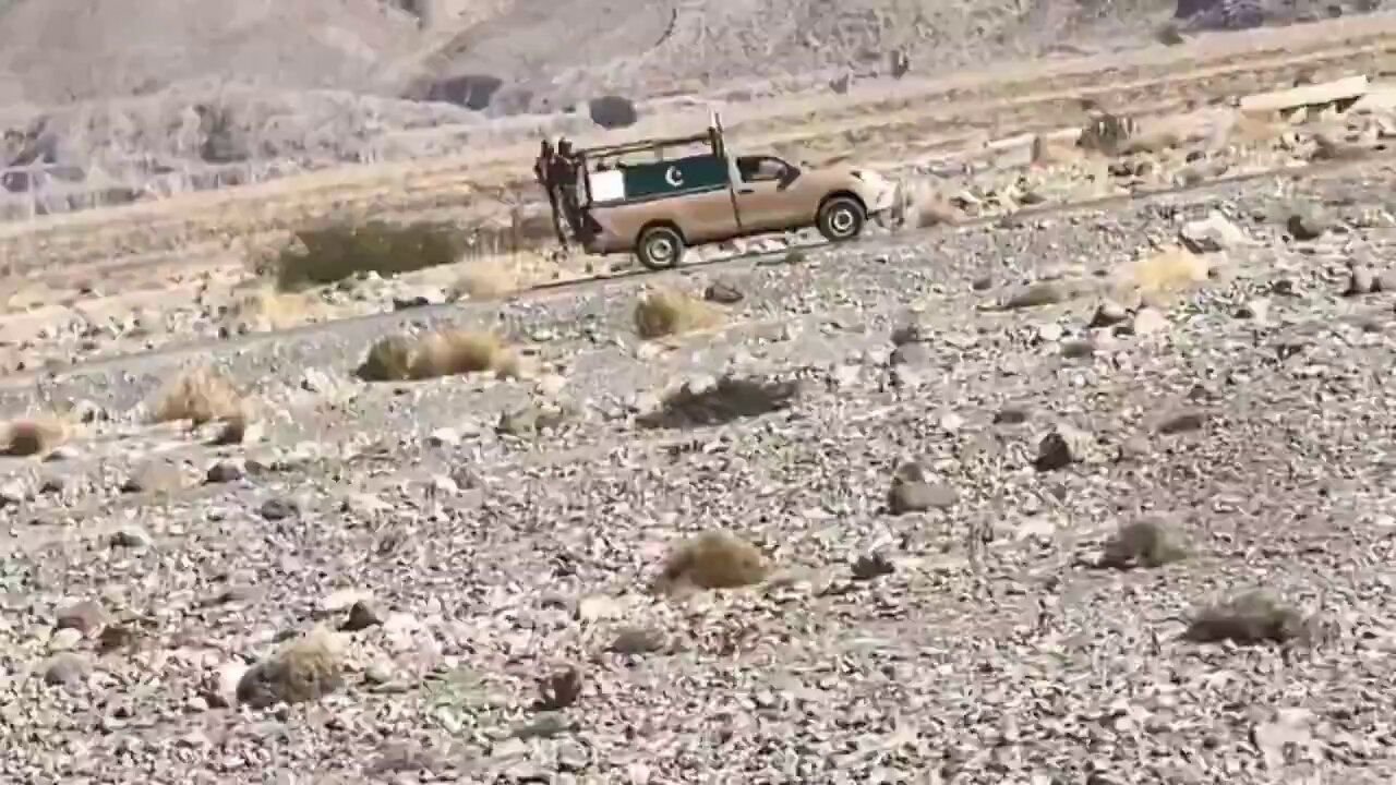 Wide shot of a rocky, arid landscape with a tan pickup truck bearing a green‑white crescent‑star flag driving on a dirt path, a person standing in the truck bed