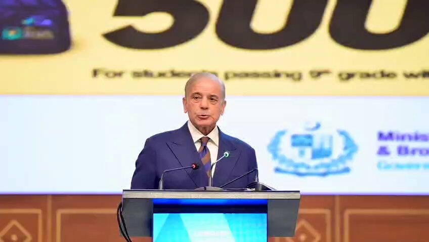 Prime Minister Shehbaz Sharif at a podium with a banner displaying the number 500 and text about ninth‑grade students, while holding a silver Chromebook