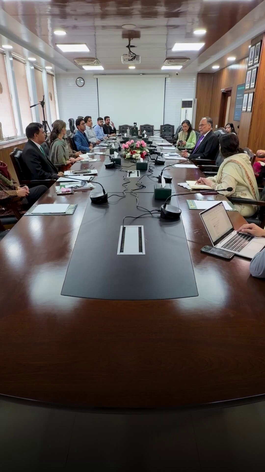 People seated around a dark wood conference table in a professional meeting room, some wearing business suits and traditional South Asian attire, with laptops and papers on the table
