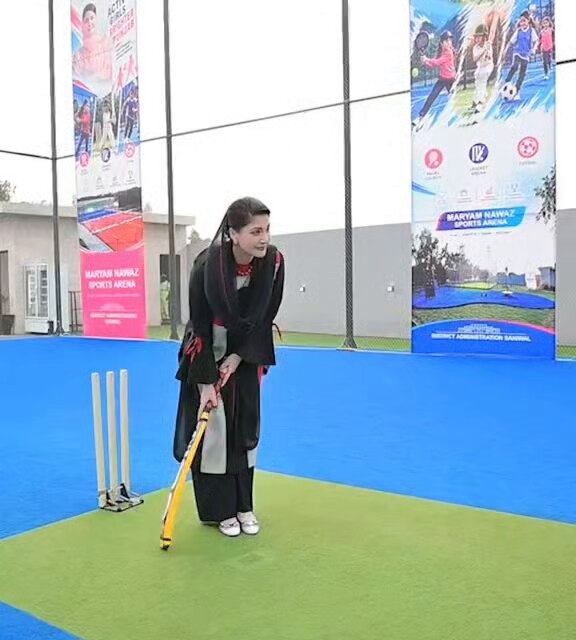 Maryam Nawaz in a black‑and‑white traditional outfit holding a yellow cricket bat on artificial turf, with banners reading “ACTIVE GIRLS” and “MARYAM NAWAZ SPORTS ARENA” behind her
