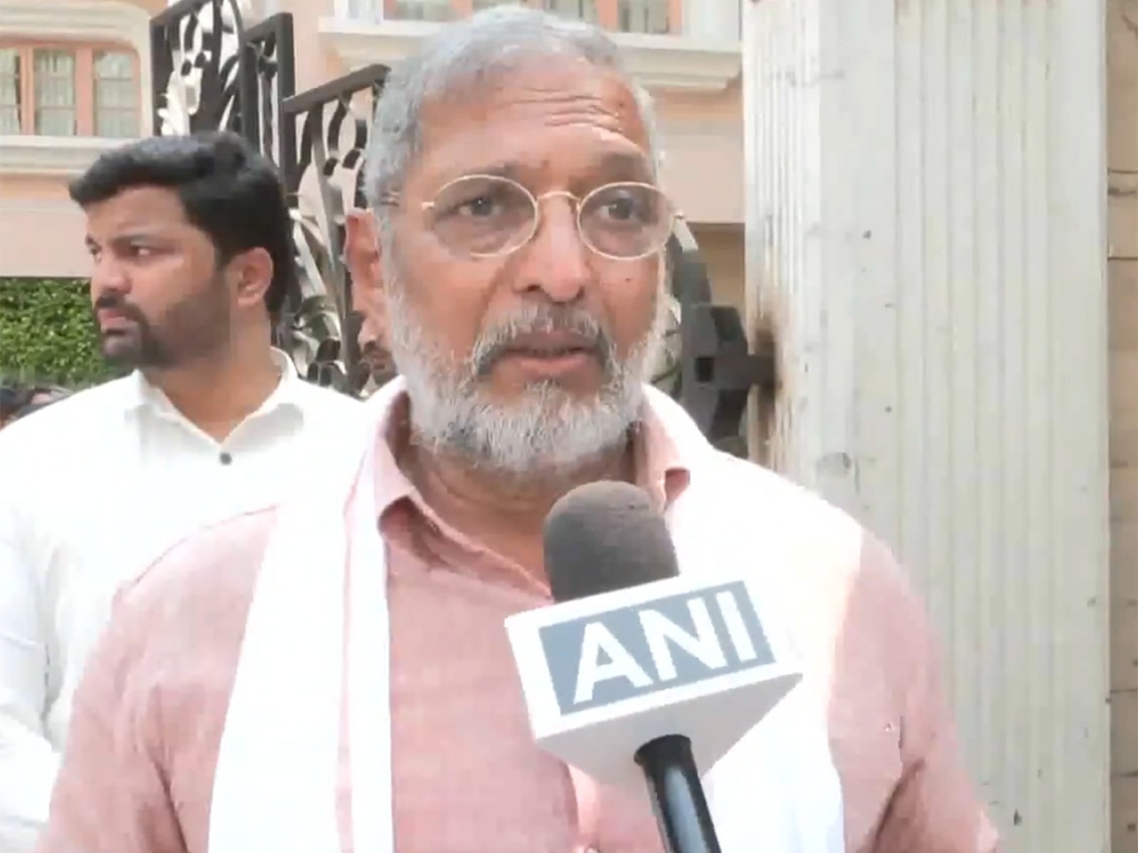 Older man with grey hair and beard, wearing glasses and a light shirt, speaking into a microphone with an ANI logo visible