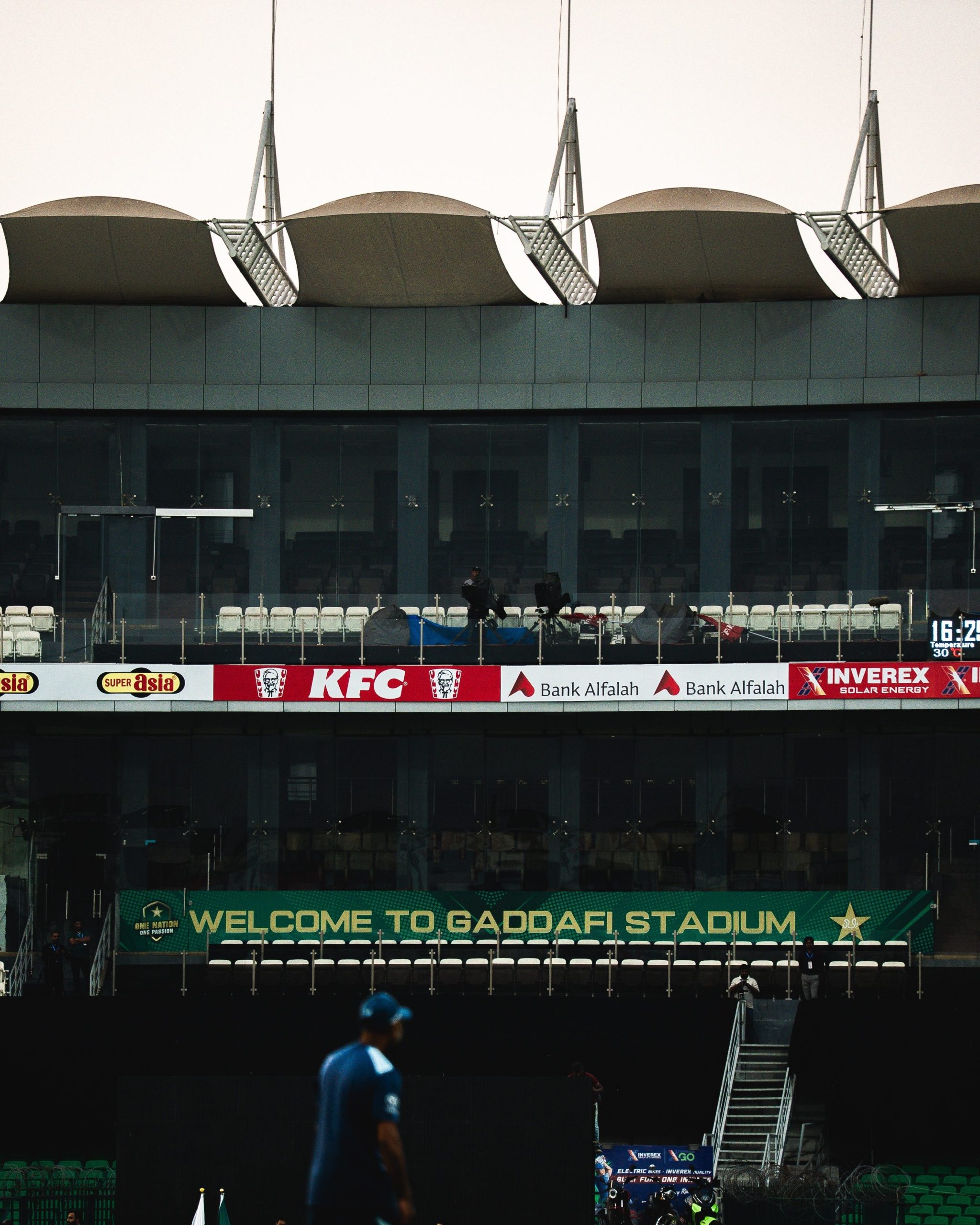 Wide shot of Gaddafi Stadium exterior featuring a modern roof, advertising banners for Super Asia, KFC, Bank Alfalah, and a sign reading 'WELCOME TO GADDAFI STADIUM' with 'ONE NATION ONE PASSION'