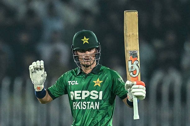 Male cricketer in a green Pakistan jersey with sponsor logos TCL and Pepsi, raising his right hand on a stadium field during a match