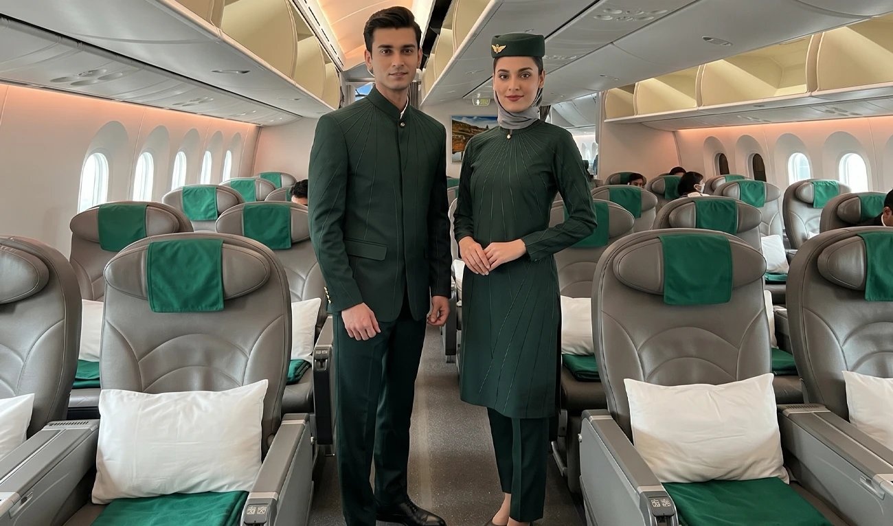 Male and female cabin crew wearing dark‑green PIA uniforms in an aircraft aisle, with grey seats and green headrest covers