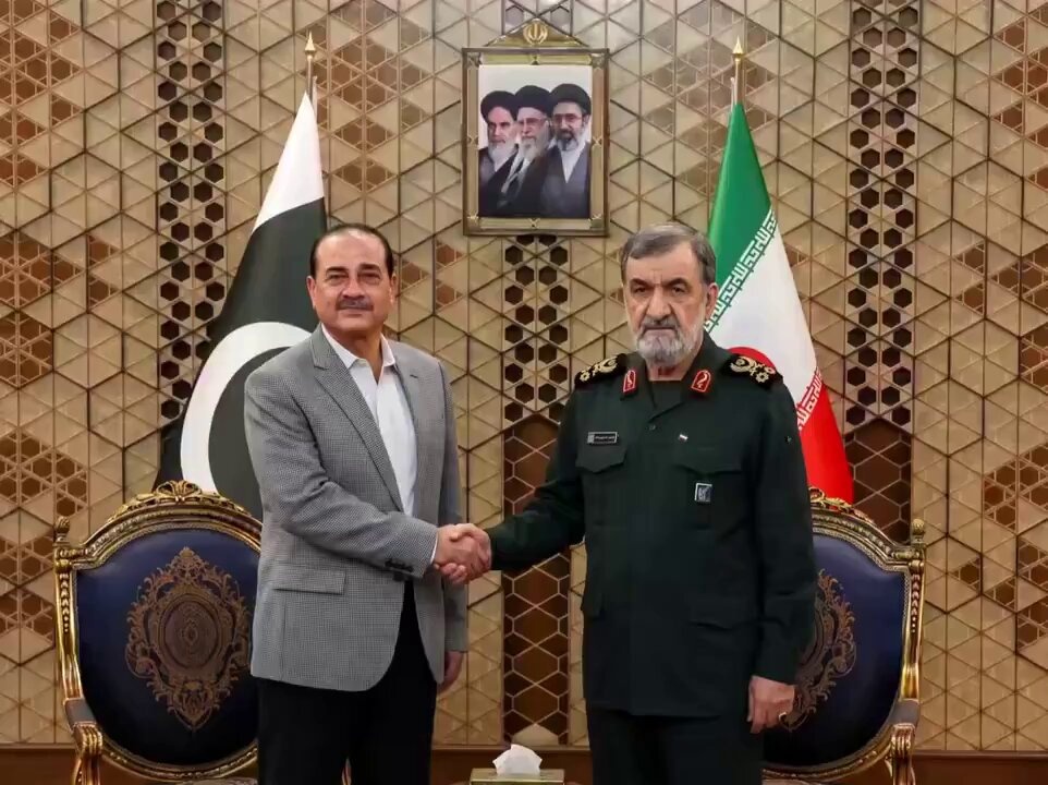 Two men shaking hands in front of a decorative wall, one in a grey blazer, the other in a dark green military uniform, with Pakistan and Iran flags displayed behind them