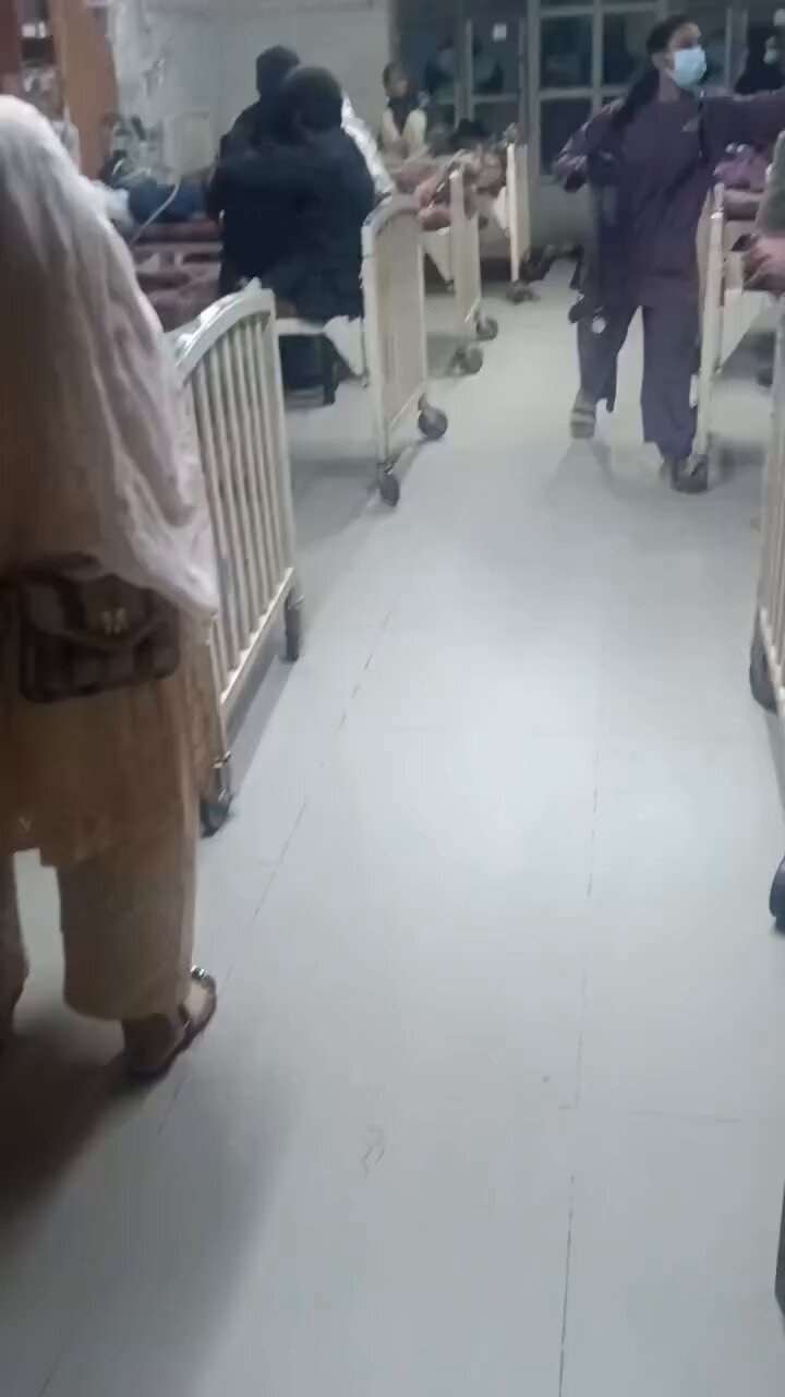 Blurred interior of a hospital ward with beds; a woman in a light‑colored headscarf and tan clothing partially visible in the foreground, and a healthcare worker in purple scrubs and a blue surgical mask walking through the room