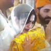 Close‑up of a woman in a white veil crying while holding a yellow patterned cloth, with a bearded man in an orange Sikh head‑covering behind her pressing his hands together
