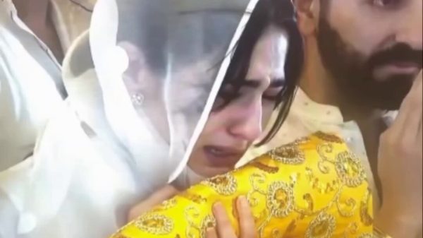 Close‑up of a woman in a white veil crying while holding a yellow patterned cloth, with a bearded man in an orange Sikh head‑covering behind her pressing his hands together