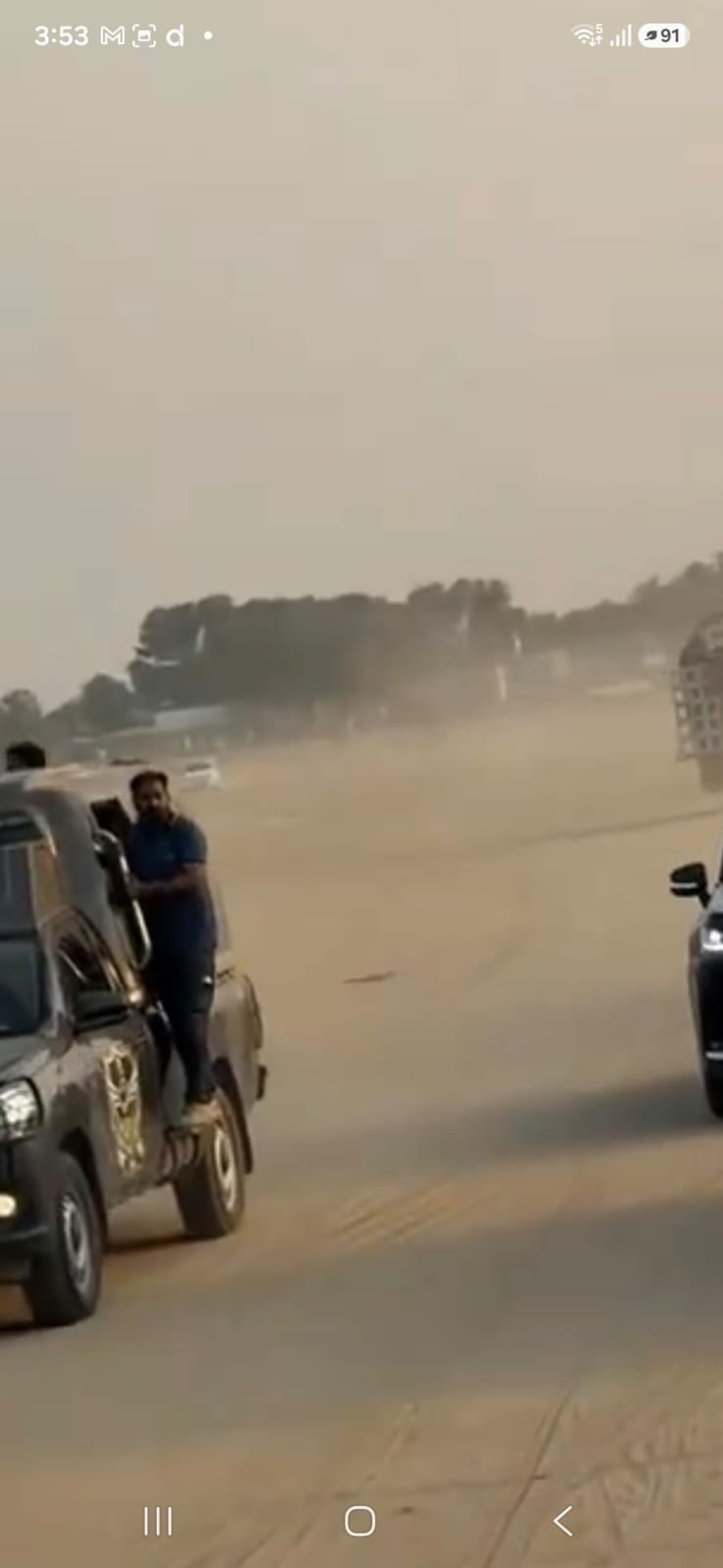 Screenshot of a mobile phone video showing a dusty desert rally scene with a man in a blue shirt beside a black SUV and another black vehicle partially visible