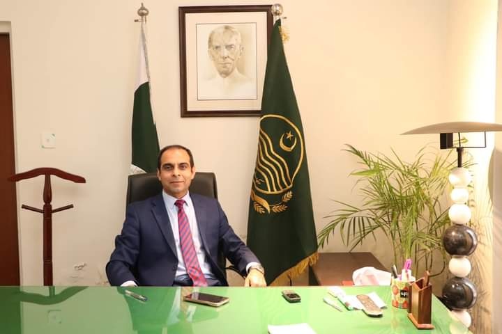 Man in dark‑blue suit seated behind a desk with Pakistan flag and a green flag, portrait of Jinnah on the wall, office décor